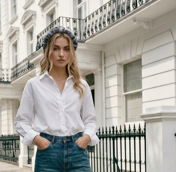 A woman stands outdoors in front of a white London-style building, wearing a white button-up shirt tucked into blue jeans and a gray headband.