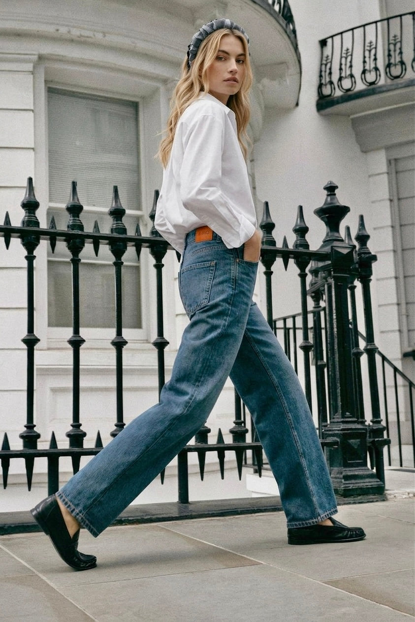 A woman walking outdoors in front of a white building with a black wrought-iron fence, wearing a grey headband, white long-sleeve blouse, wide-leg blue jeans, and black loafers.