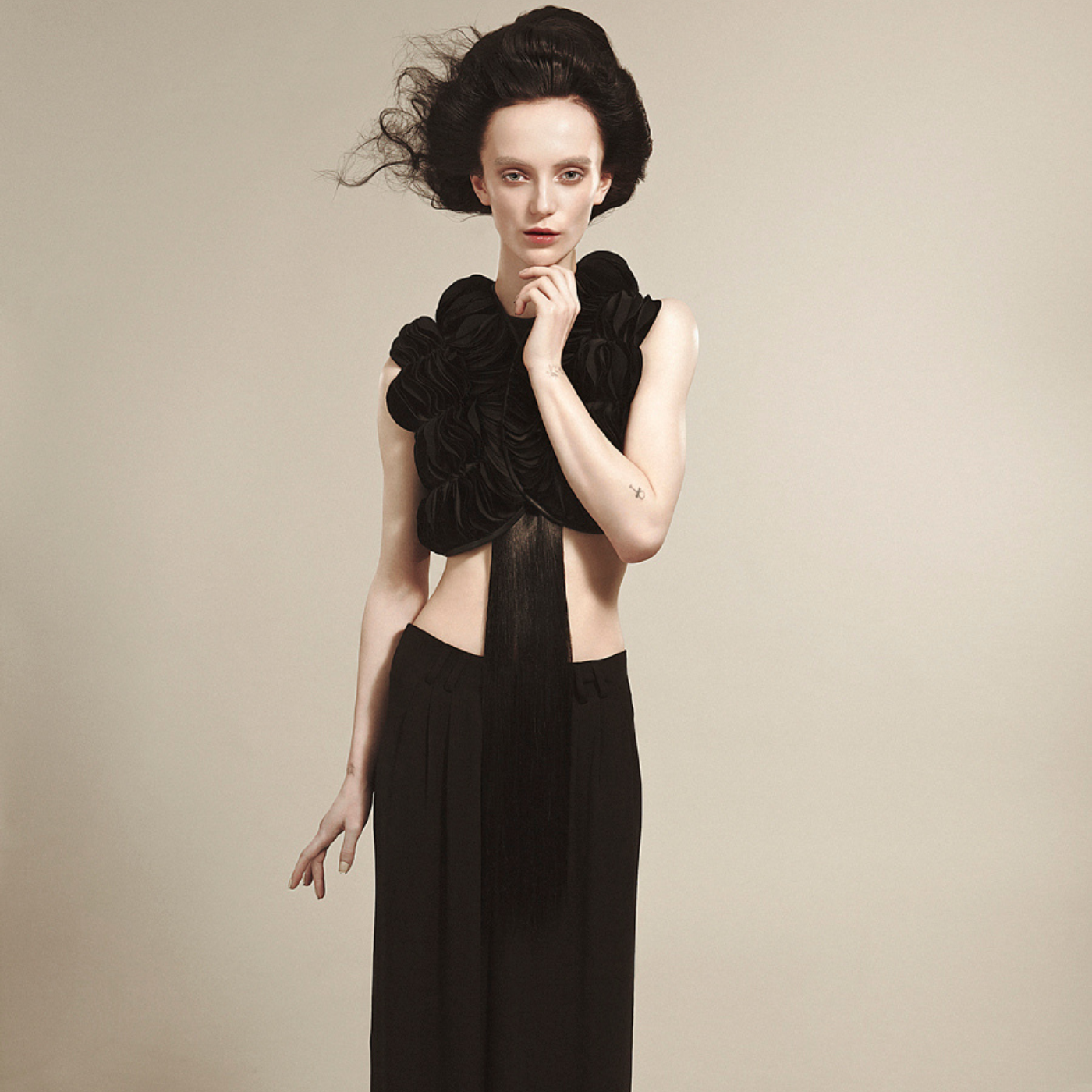 Model wearing a black sleeveless top with an intricate design with hair and high-waisted black pants on a beige background.