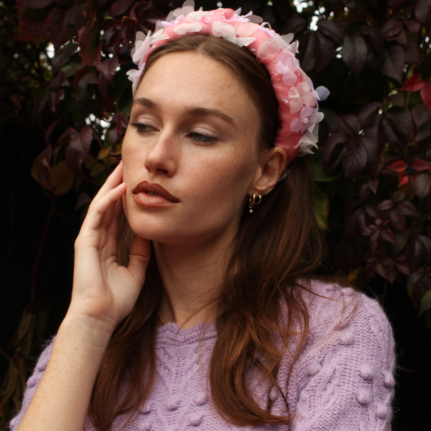 Woman wearing a pink floral headband and purple sweater against a dark leafy background