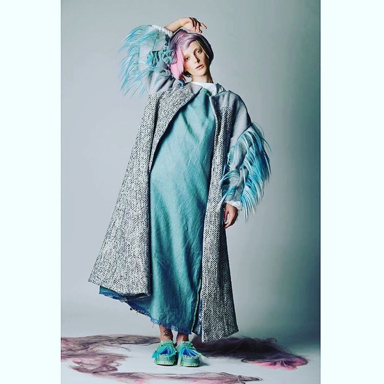 Model wearing a teal dress with a patterned shawl and colorful headpiece on a plain background