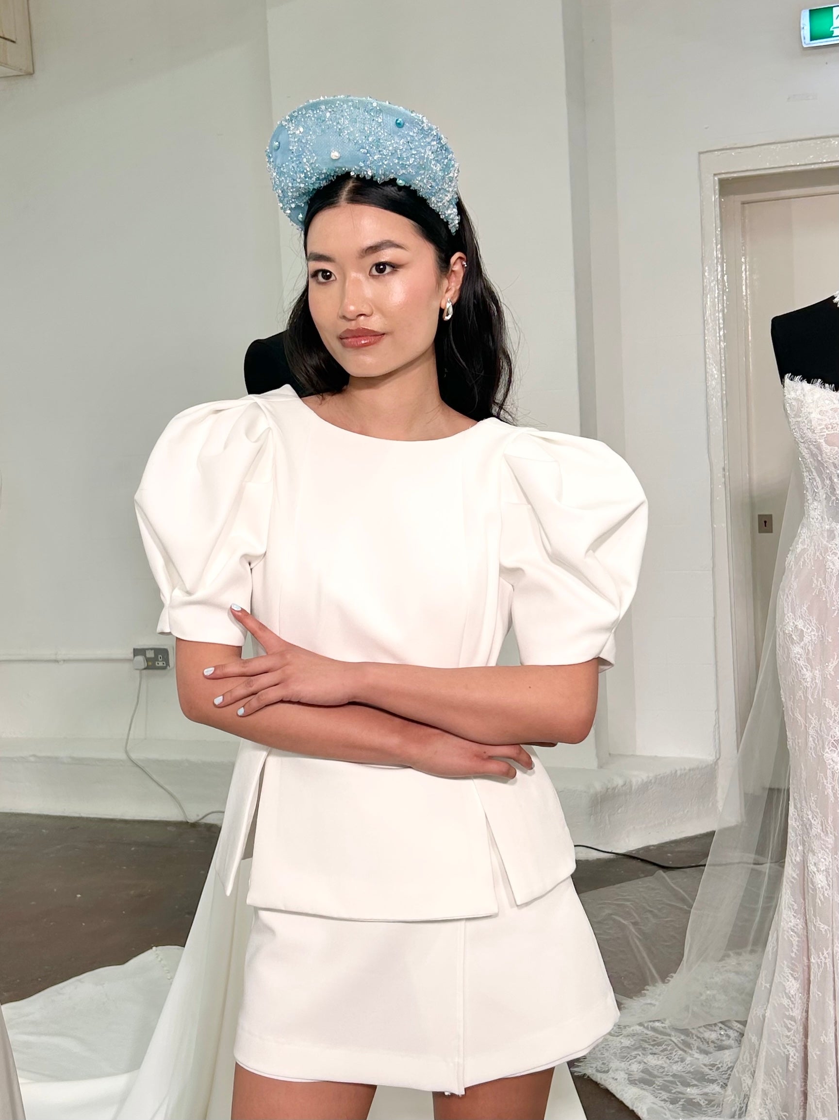Model wearing a white outfit with puffed sleeves and a headband, standing in a room with white walls and a door.