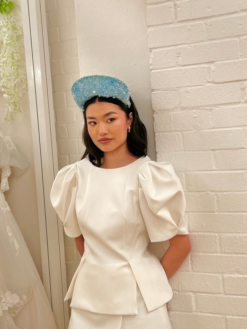 Model wearing a white outfit with puffed sleeves against a white brick wall.