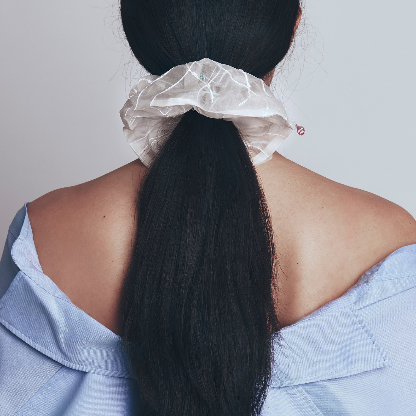 Model with long black hair tied in a ponytail with a white scrunchie, wearing a light blue off-shoulder top.