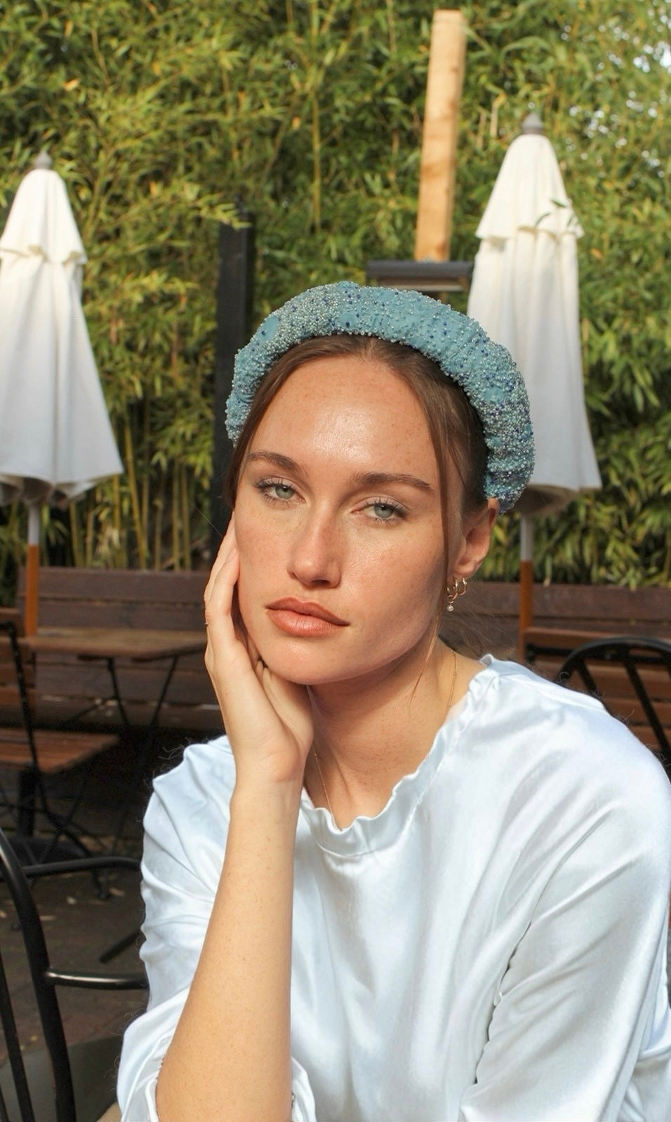Woman wearing a light blue headband outdoors with greenery and white umbrellas in the background