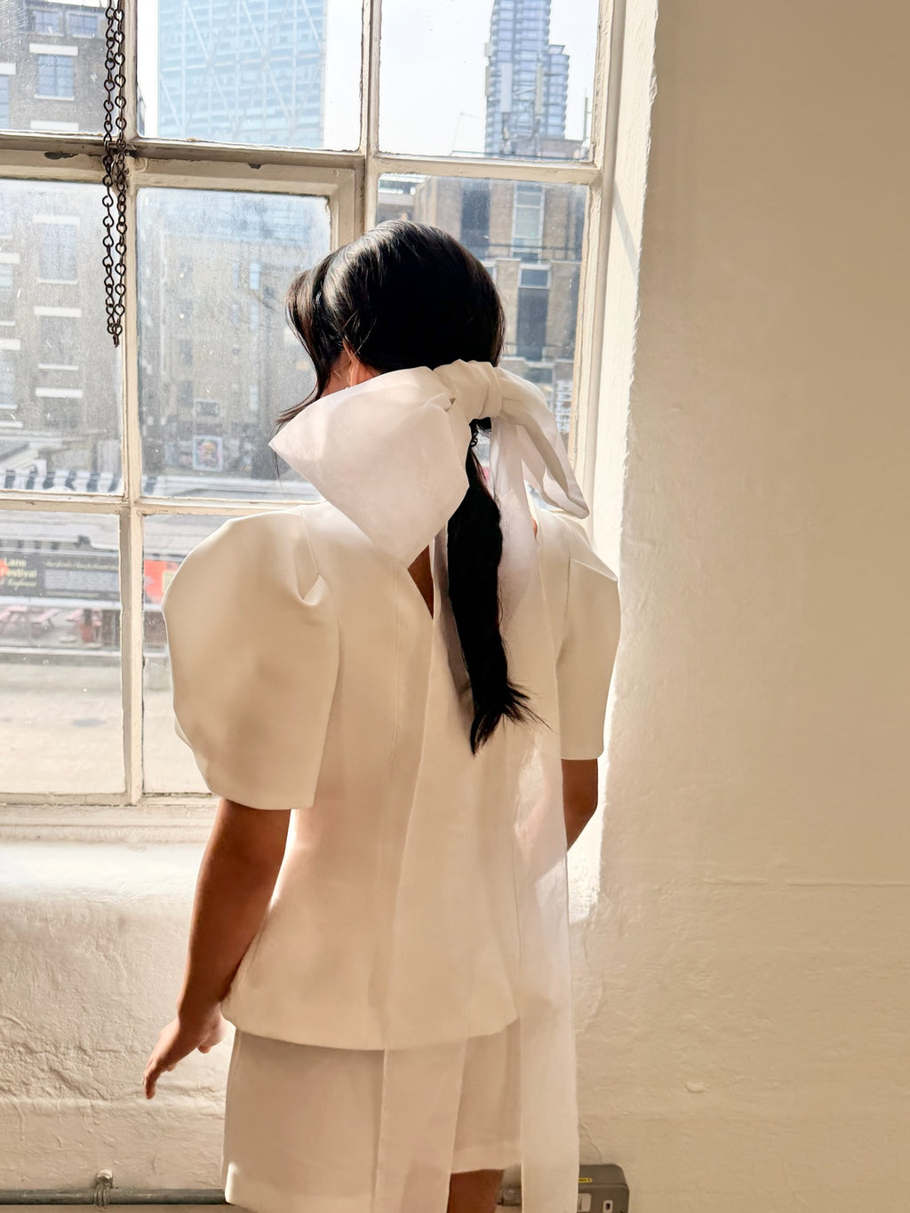 Model wearing a white outfit with a unique bow design, standing in front of a window with cityscape view.