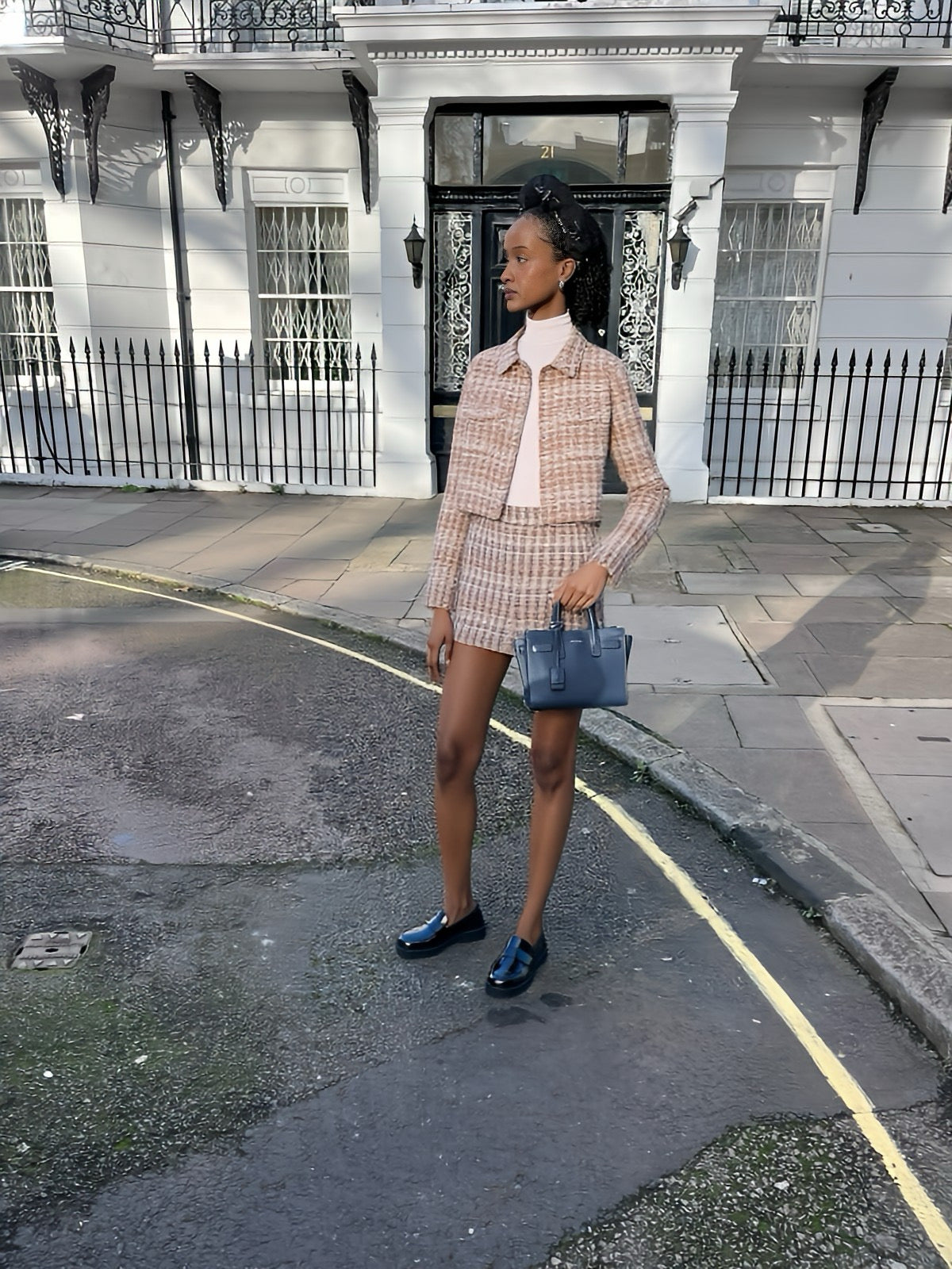 Woman in a plaid outfit standing on a street with a white building in the background