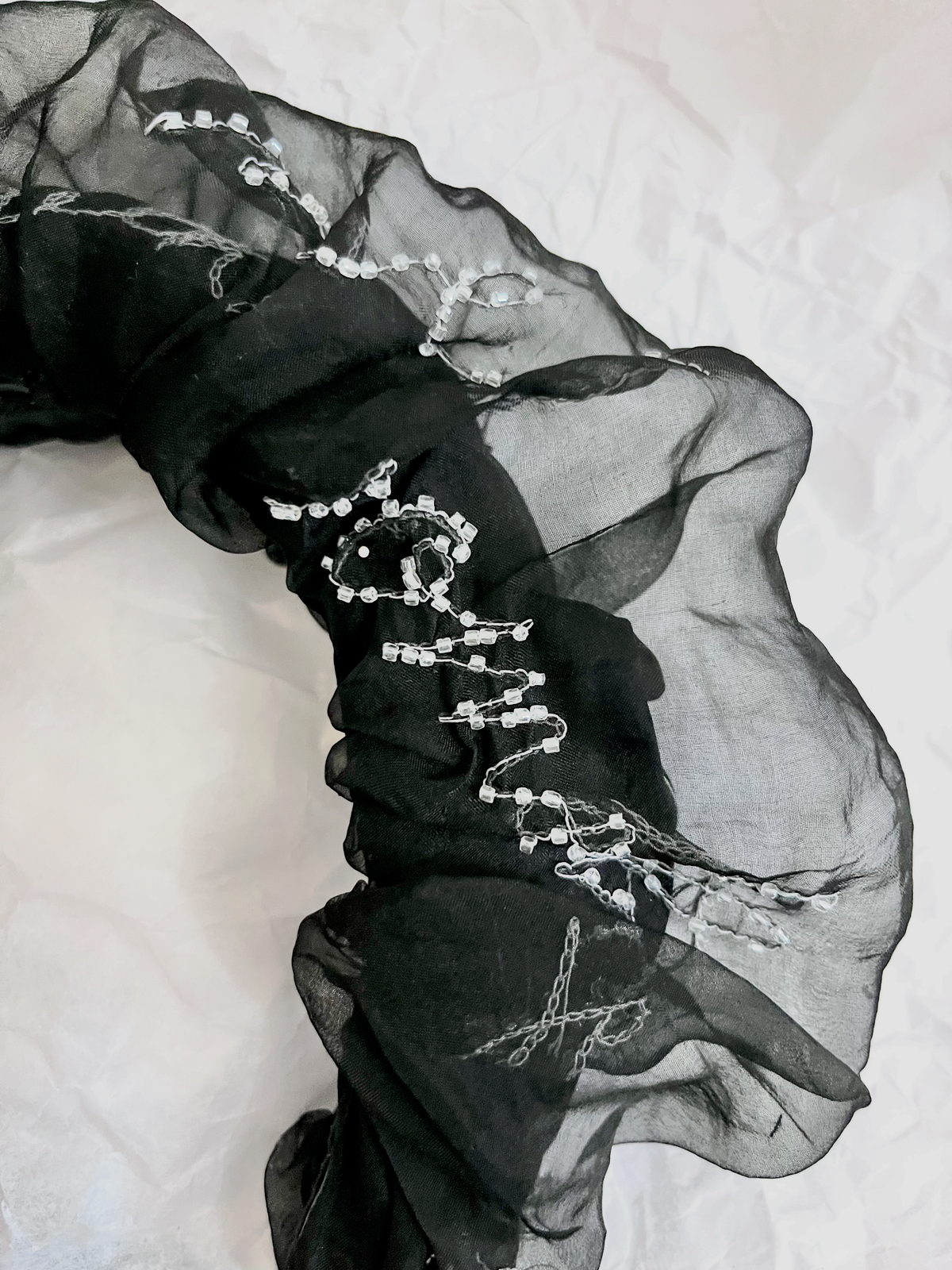 A sheer black organza hair accessory,  featuring intricate silver bead and chain embroidery, laid out on a white textured fabric background.