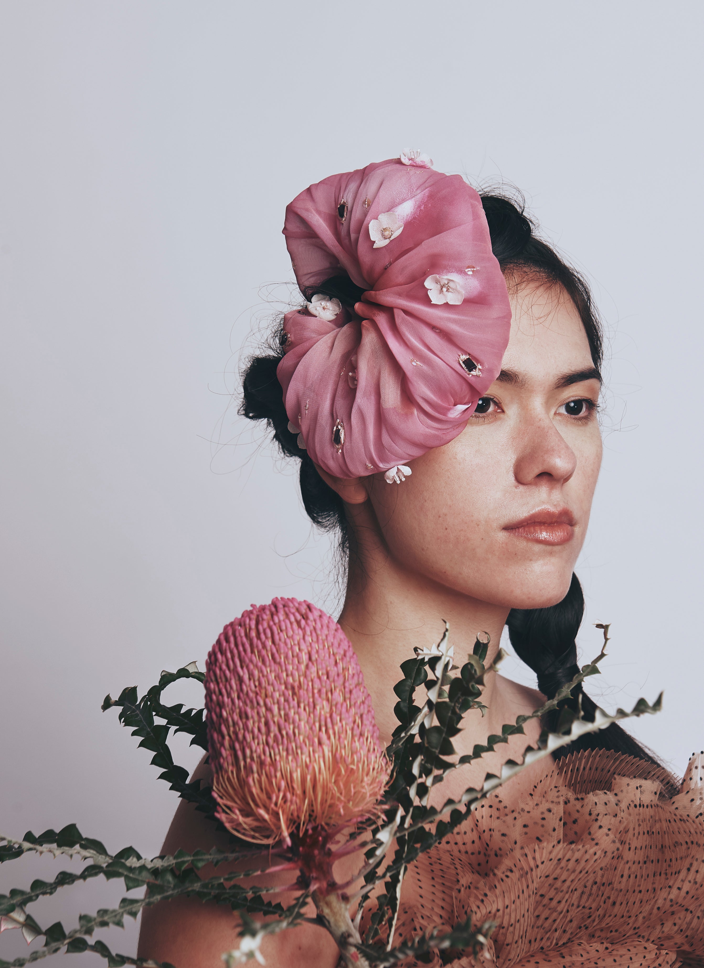 Woman wearing a pink scrunchy with floral details, surrounded by pink flowers and green leaves on a light gray background.