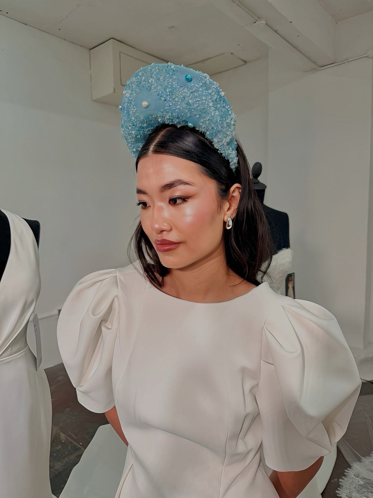 Model wearing a white dress with puffed sleeves and a blue decorative headpiece in an indoor setting.