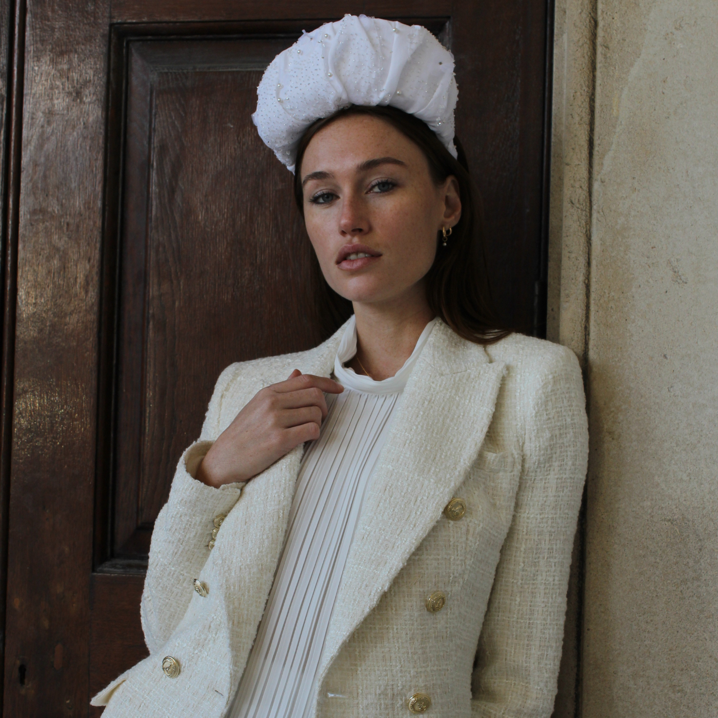 Model wearing a white coat and headband against a textured wall and wooden door.