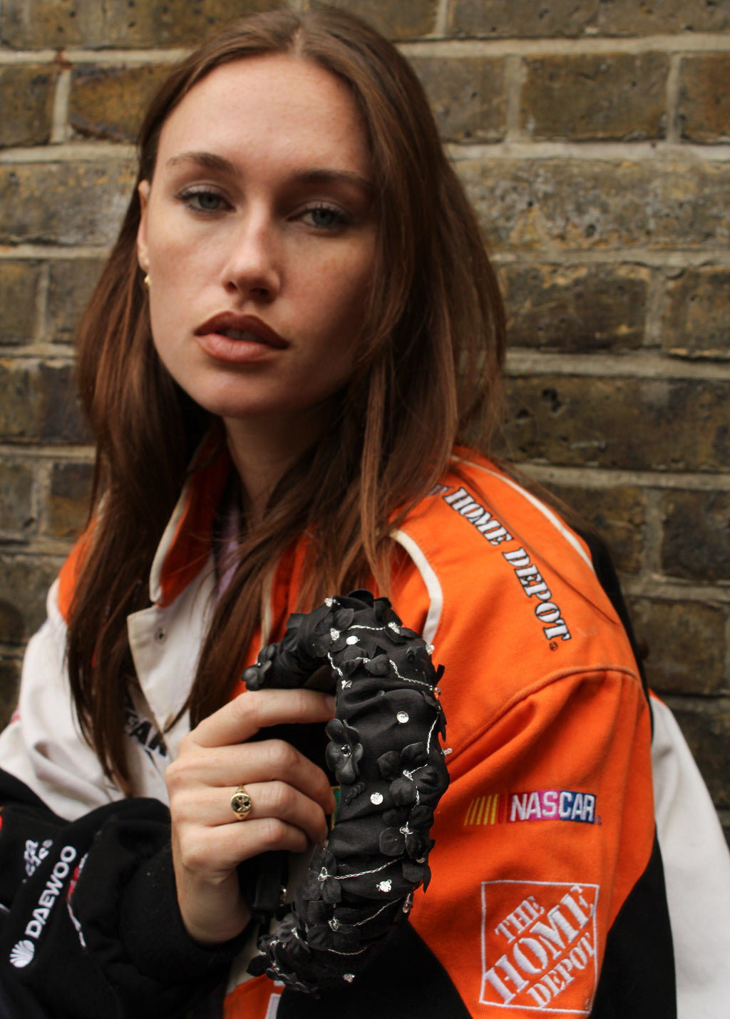 Model wearing an orange and white jacket with visible branding, holding a black headband against a brick wall.